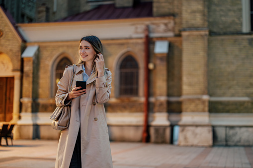 Portrait of a beautiful young stylish female walking in the city streets and holding a mobile phone in her hands. Fashionable woman in trench coat smiling while typing a message on her smartphone.