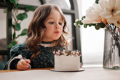 Child birthday party. Adorable little girl sits by the table with birthday cake decorated flowers and burn candle, makes wish. Kid in festive dress at room with balloons, bouquet of flowers at vase.