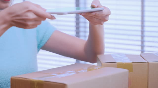 Young woman small business owner using mobile app checking parcel box. Warehouse worker, seller holding phone scanning retail drop shipping package postal parcel on cell preparing ship order.
