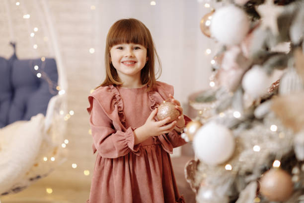 маленькая очаровательная девочка в нежно-розовом платье вешает золотой блестящий шар на рождественскую елку с подсветкой. улыбающийся реб - happy new years стоковые фото и изображения