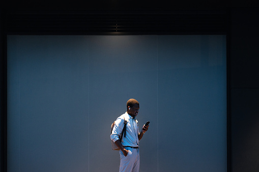 Handsome African-American business man listening to music and typing text messages on his smartphone while standing in front of a blue wall outdoors.