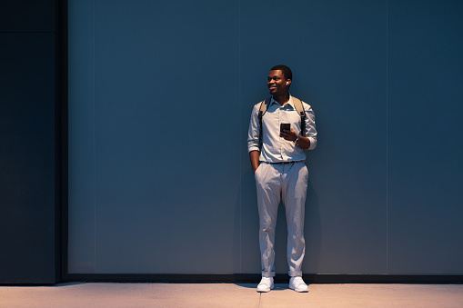 Smiling African-American business man listening to music and typing text messages on his smartphone while standing in front of a blue wall and looking away.
