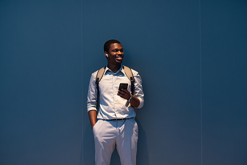 Happy African-American business man listening to music and typing text messages on his smartphone while standing in front of a blue wall and looking away.