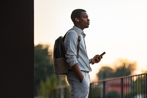 Handsome African-American business man with a backpack listening to music and typing text messages on his smartphone while standing on a balcony.