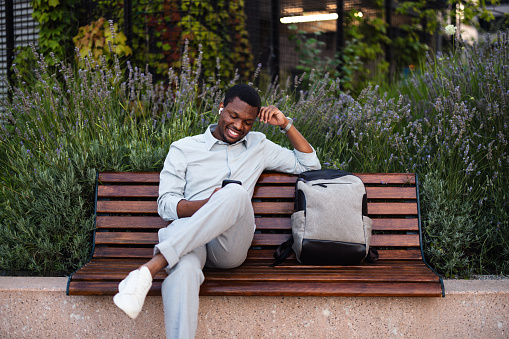 Happy African-American business man listening to music and typing text messages on his smartphone while sitting on a bench with his backpack.