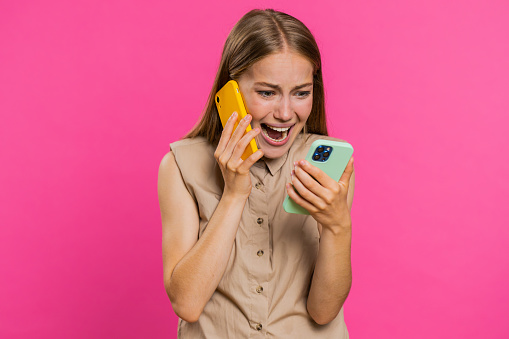 Irritated nervous stressed young woman talking screaming on two mobile phones having conversation conflict quarrel complaint dispute discuss solve problem. Girl isolated on pink studio background