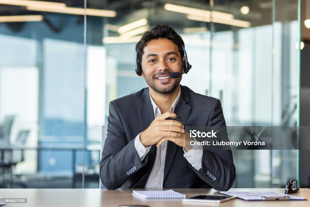 Close-up portrait of a young Indian man sitting in the office at the desk wearing a headset, smiling and talking to the camera Close-up portrait of a young Indian man sitting in the office at the desk wearing a headset, smiling and talking to the camera. Latin American and Hispanic Ethnicity Stock Photo