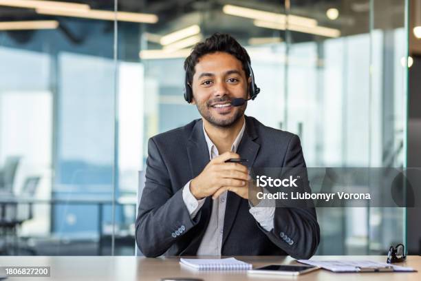 Closeup Portrait Of A Young Indian Man Sitting In The Office At The Desk Wearing A Headset Smiling And Talking To The Camera Stock Photo - Download Image Now