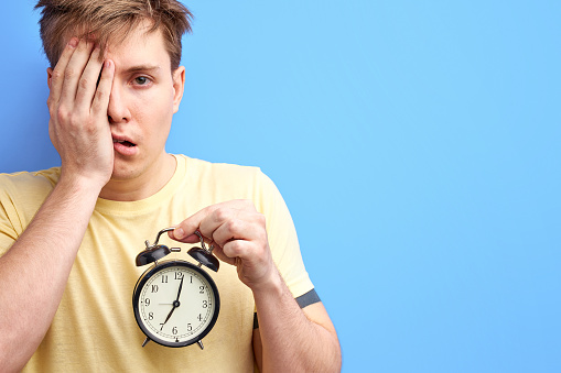 student want to sleep before university, stand closing half of face with hand, looking at camera and holding alarm- clock in hands. isolated blue background