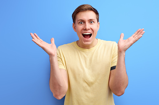 man rejoices and celebrating winning on blue background, positive open-minded male gesturing, raising hands and looking at camera with opened mouth, get good news