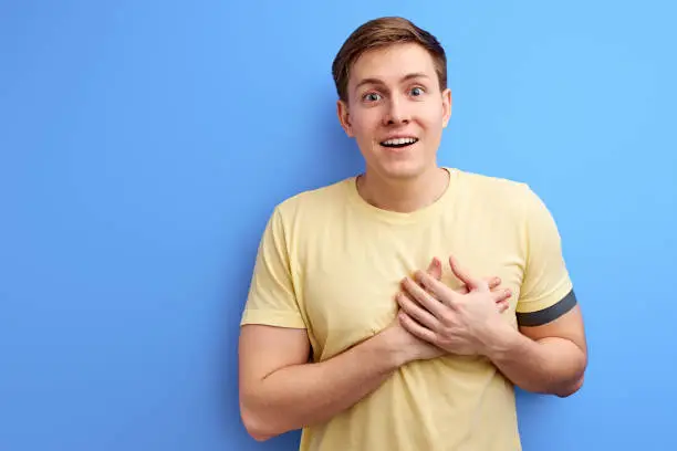 pleasant happyyoung man holding hands on chest posing isolated on blue wall background. male is being cordial and friendly, expresses gratitude and thankfulness, has pleasant smile