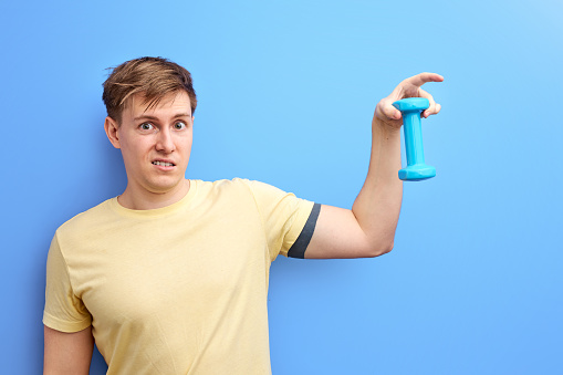 sportsman holding small dumbbell, it is not difficult for him to raise. isolated blue background. studio portrait of caucasian guy in casual t-shirt. sport concept