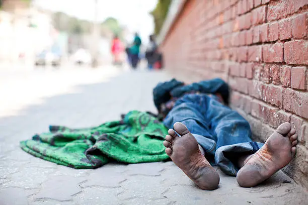 Homeless man lies on the sidewalk. In the focus on foreground are his dirty feet. Photographed on street in Kathmadu