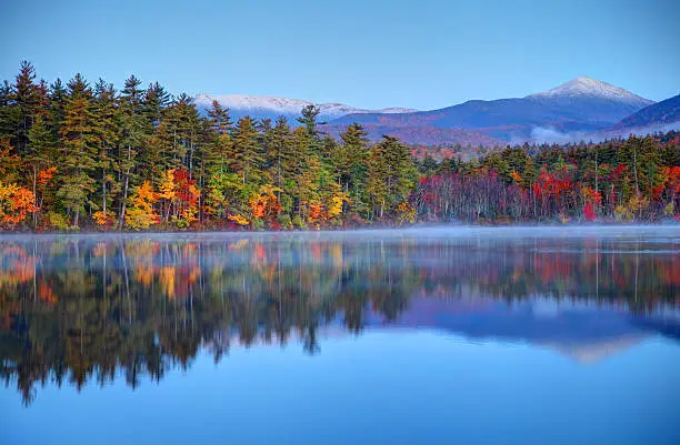 Autumn snowcapped mountains in New Hampshire. Photo taken at dawn on a calm lake during the peak fall foliage season near the White Mountains National Forest. New Hampshire is one of New England's most popular fall foliage destinations bringing out some of  the best foliage in the United States