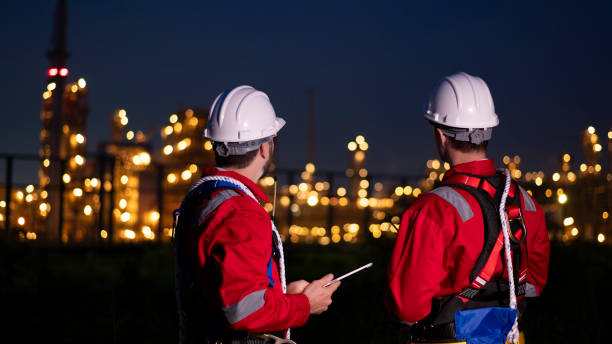 istock Engineer survey team wear uniform and helmet stand workplace checking blueprint project and radio communication inspection work construction site with night lights oil refinery background. 1807046542