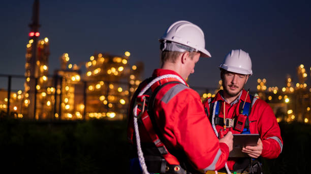 istock Engineer survey team wear uniform and helmet stand workplace checking blueprint project and radio communication inspection work construction site with night lights oil refinery background. 1807046570