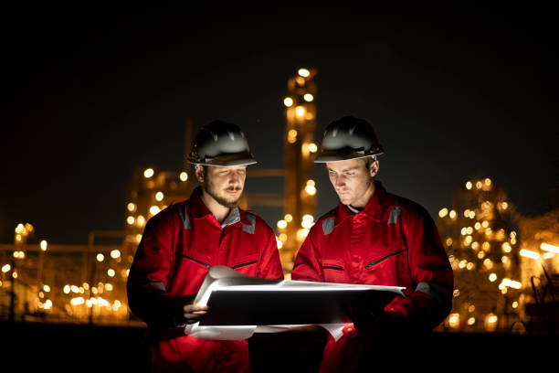 istock Engineer survey team wear uniform and helmet stand workplace checking blueprint project and radio communication inspection work construction site with night lights oil refinery background. 1807046627