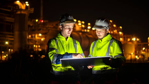 istock Engineer survey team wear uniform and helmet stand workplace checking blueprint project and radio communication inspection work construction site with night lights oil refinery background. 1807046715