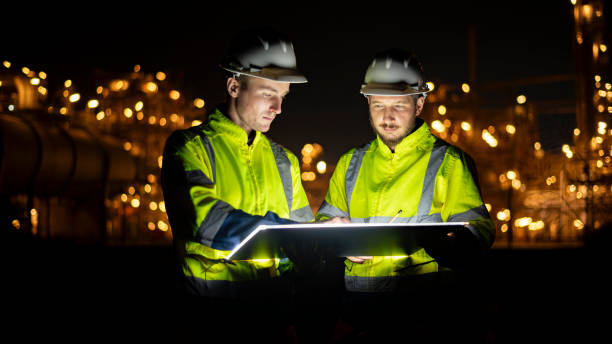 istock Engineer survey team wear uniform and helmet stand workplace checking blueprint project and radio communication inspection work construction site with night lights oil refinery background. 1807046729