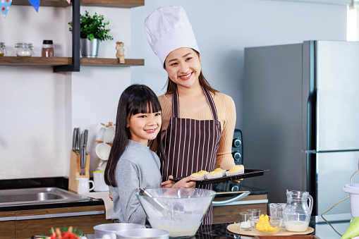 Asian female baker pastry chef mother wears white tall hat and apron standing smiling with little girl daughter holding showing presenting delicious tasty homemade baking bakery cupcakes in tray.