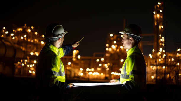 istock Engineer survey team wear uniform and helmet stand workplace checking blueprint project and radio communication inspection work construction site with night lights oil refinery background. 1807046776