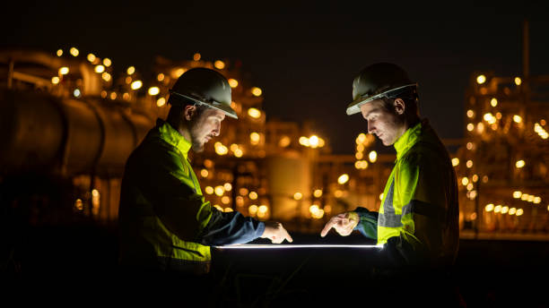 istock Engineer survey team wear uniform and helmet stand workplace checking blueprint project and radio communication inspection work construction site with night lights oil refinery background. 1807046782