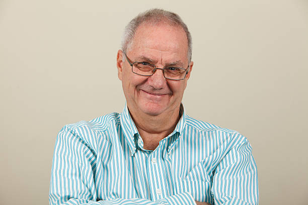 Portrait of balding senior man wearing bifocal glasses, vest and plaid shirt, dark background with window light illuminating one side of his face.