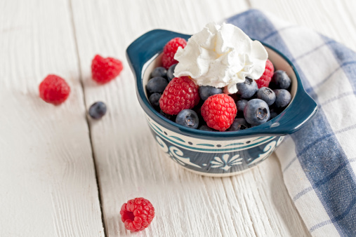 A bowl of raspberries and blueberries on white wood with a blue and white plaid napkin.A bowl of raspberries and blueberries on white wood with a blue and white plaid napkin and whipped cream.
