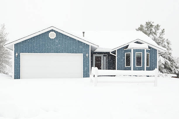 A small quaint house during a winter snowstorm. A large amount of snow has accumulated on the house and snowflakes are visible against the dark areas of the image. This would make a great home security concept image as an unshoveled house is instant notification no one is at home.