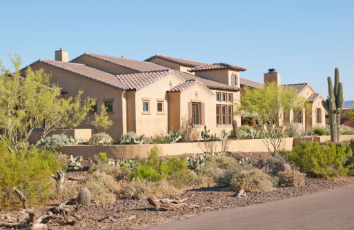Arizona desert home in Pueblo or Santa Fe style.  Parapet roof with visible drain pipes protruding through.  Desert landscaping including saguaro and other cacti.  Courtyard with low fence and arched entry.