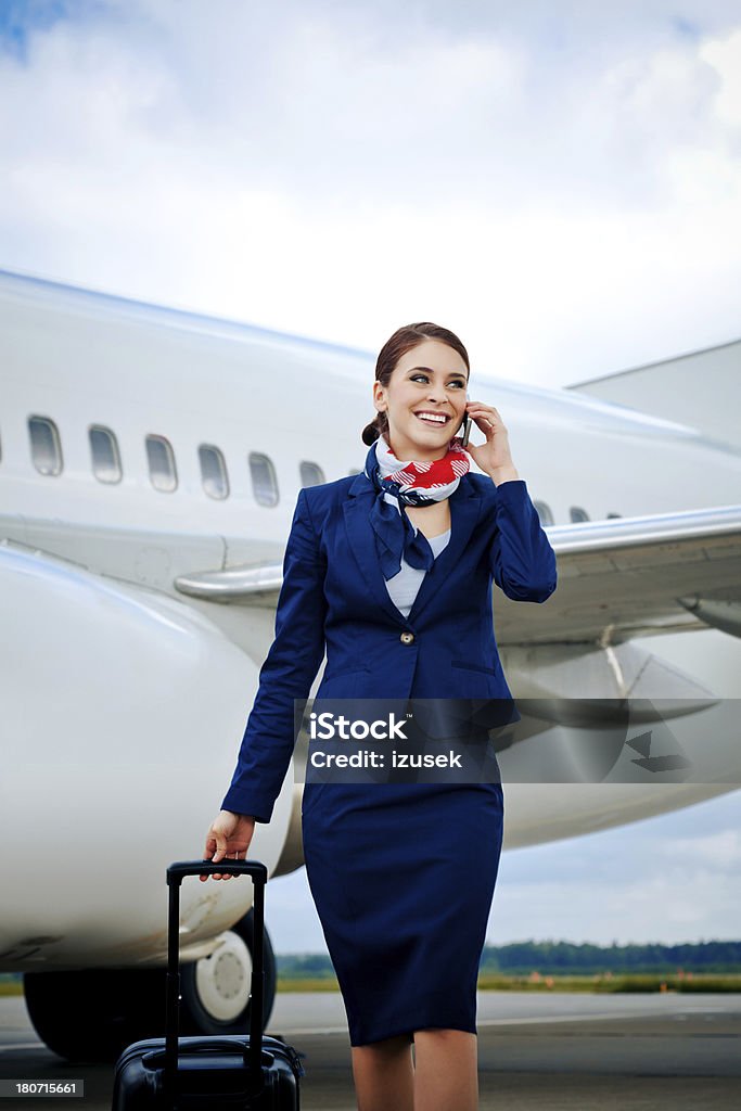 Air stewardess talking on phone Outdoor portrait of a cheerful flight attendant walking with the suitcase with an aircraft in the background and talking on mobile phone. Cabin Crew Stock Photo