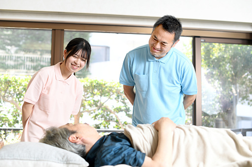 A middle-aged Asian male caregiver and a young female caregiver talk to a bedridden elderly man.