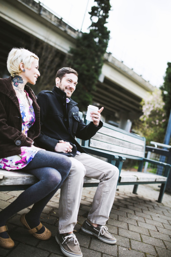 A well dressed young couple sit on a bench outside on the Willamette river waterfront in Portland, Oregon, smiling at the camera.  The man holds a coffee cup.  Vertical with copy space.