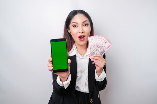 Surprised Asian businesswoman wearing black suit showing her smartphone and money in Indonesian Rupiah, isolated by white background