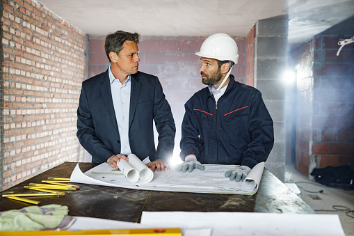 Male inspector and manual worker communicating while examining blueprints at construction site.