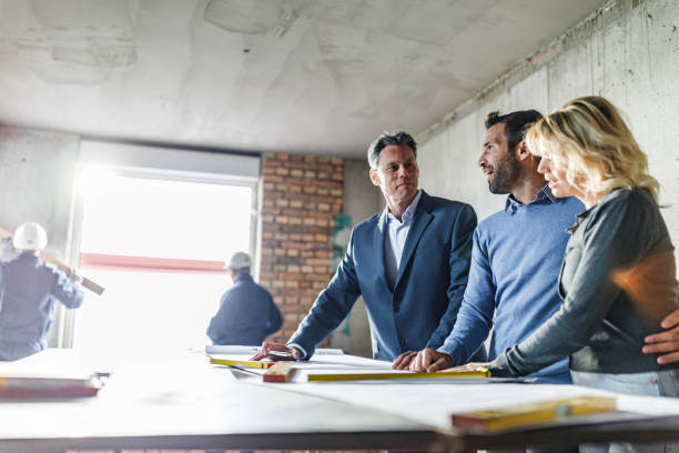 istock Young couple and their agent talking while examining blueprints at construction site. 1807259108