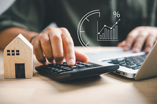 Hand using calculators, planning home refinance. Wooden house model, buy or rent note, calculators on desk. Saving for property purchase, mortgage payment. Tax, credit analysis for financial benefit.