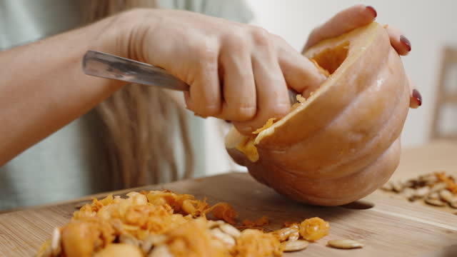 Approaching a Young Woman with Long Hair Who is Carving a Pumpkin for Dinner. White Interior and Wooden Table.
