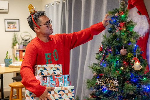 An asian male holds gifts next to christmas tree
