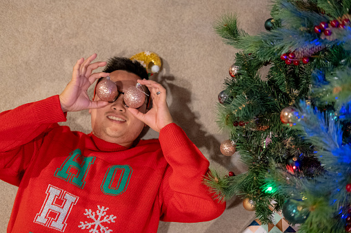Young asian male lying next to christmas tree holding christmas balls to eyes