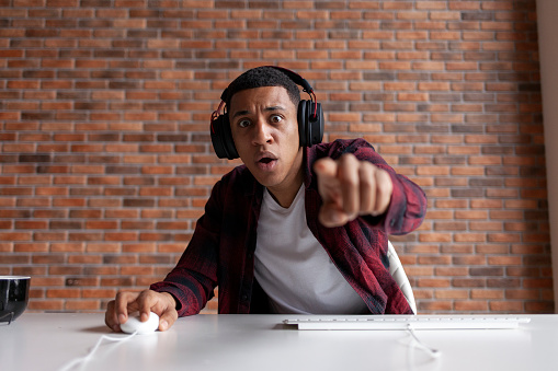 young surprised guy african american gamer plays video game on computer at home and points finger at the screen, shocked man in headphones sits at table and points to monitor