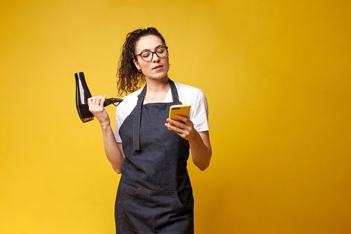 young girl hairdresser in uniform holds hair dryer and uses smartphone on yellow background, woman stylist in apron holds phone and types