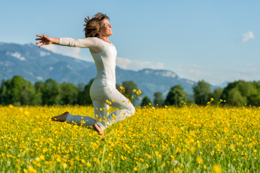 smiling mature woman jumping barefoot in flower meadow with arms outstretched