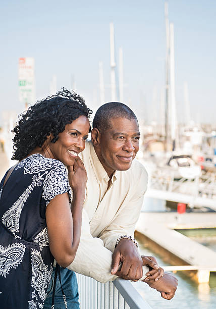 Older couple smiling at harbor Senior couple of African descent comfortably leaning on rail smiling at harbor with the boat docks in the background. lypsela2013 stock pictures, royalty-free photos & images