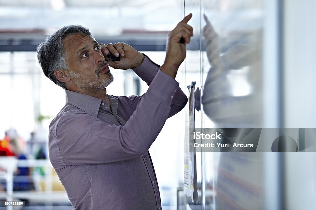 I'm looking at the figures right now A mature businessman talking on his cellphone while looking at something on the board Adult Stock Photo