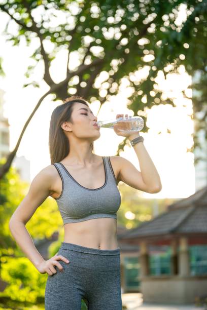 junge asiatische frau in sportbekleidung mit smartwatch, die nach dem training in einem öffentlichen park feuchtigkeit und entspannung genießt und einen fitten körper im warmen sonnenuntergang präsentiert. - exercising sensuality water bottle relaxation exercise stock-fotos und bilder