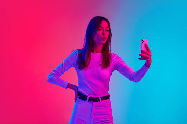 istock Young beautiful woman taking selfie with mobile phone against gradient pink blue studio background in neon light. Social media 1807390799