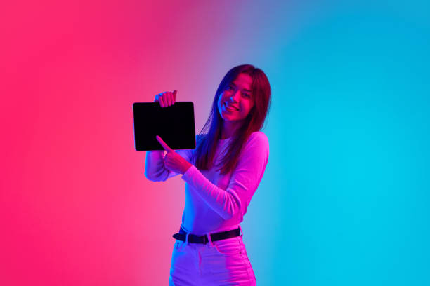 istock Young smiling woman showing tablet screen against gradient pink blue studio background in neon light 1807390862