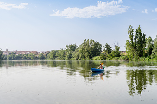 On the Duero River as it passes through Tordesillas, Valladolid-Spain, a middle-aged man with no hair or shirt is in the calm waters of the river with a recreational canoe. Weekend recreational activity. The river banks have abundant green vegetation.