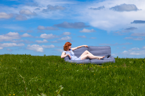 Redhead in a blue dress reclining on her sofa in the middle of a field in Summer.